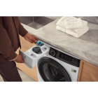 Valkoinen etupäästä täytettävä pyykinpesukone, jossa on avoin pesuainelokero, harmaa ohjauspaneeli ja lasiovi. Se on sijoitettu puunväristen kaappien väliin, ja päällä on taiteltuja pyyhkeitä.